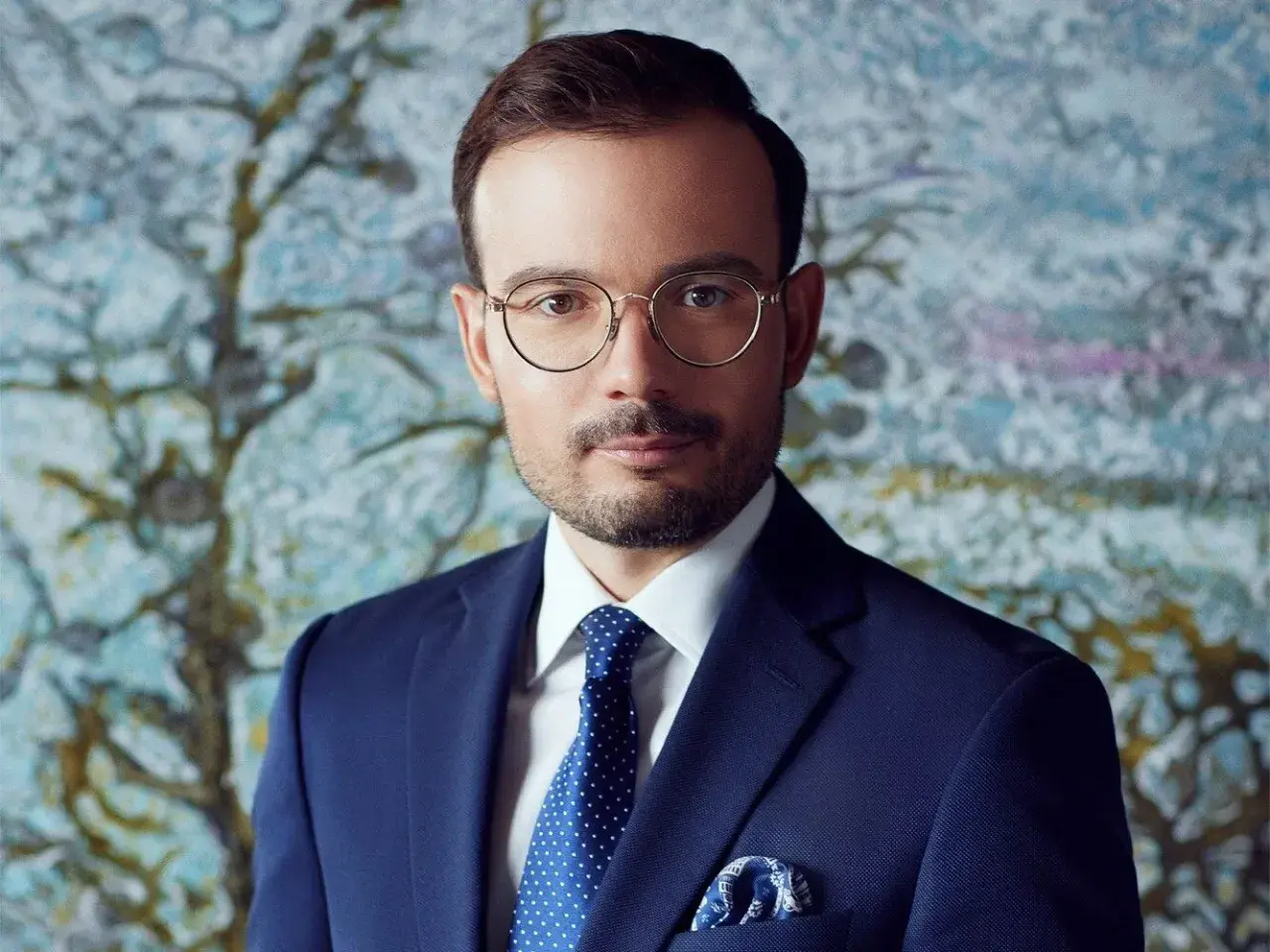 Mężczyzna w granatowym garniturze, białej koszuli i niebieskim krawacie w kropki, w okularach, na tle abstrakcyjnego obrazu.
