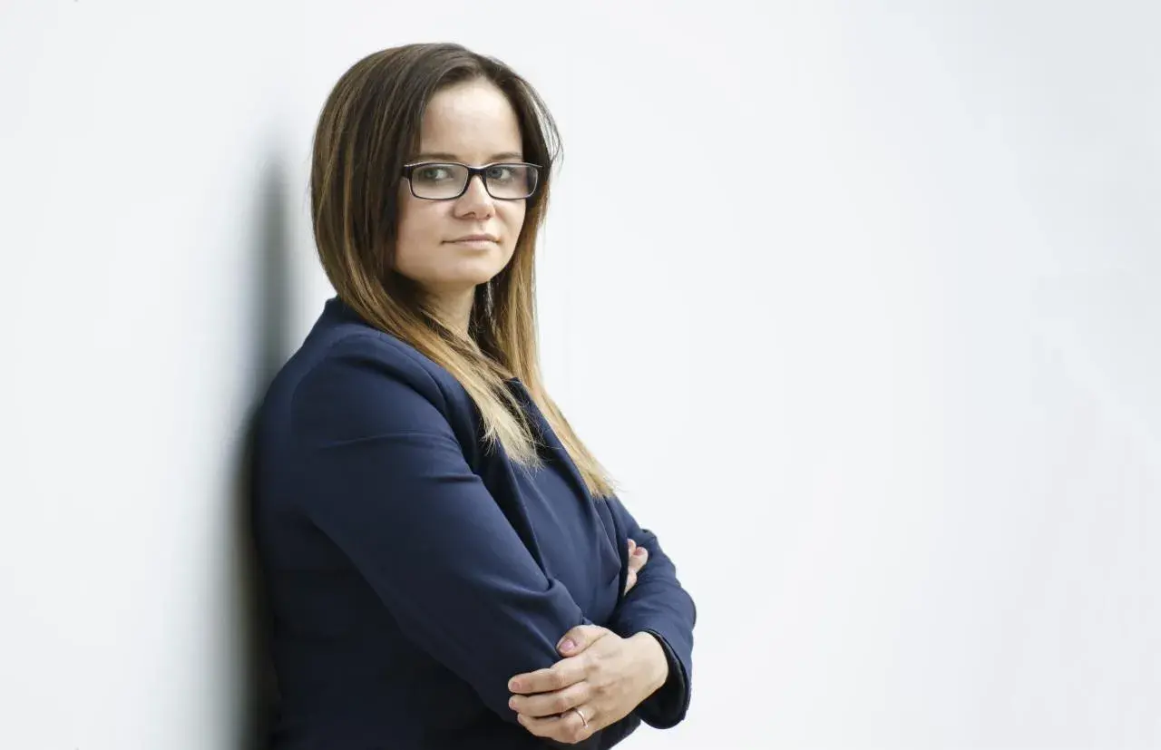 Kobieta w okularach i granatowej marynarce stoi oparta o ścianę, ze skrzyżowanymi rękami.