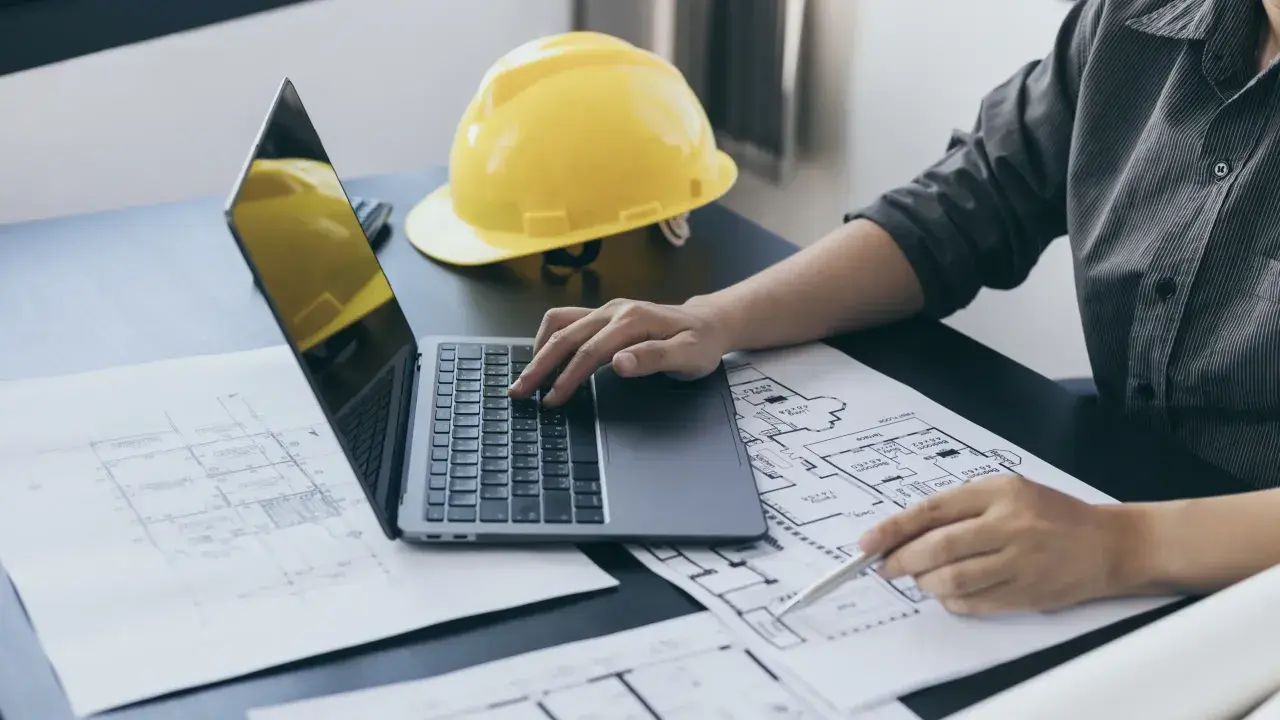 Inżynier analizuje plany budowlane, obliczając powierzchnię zabudowy na laptopie. Obok leży żółty kask.