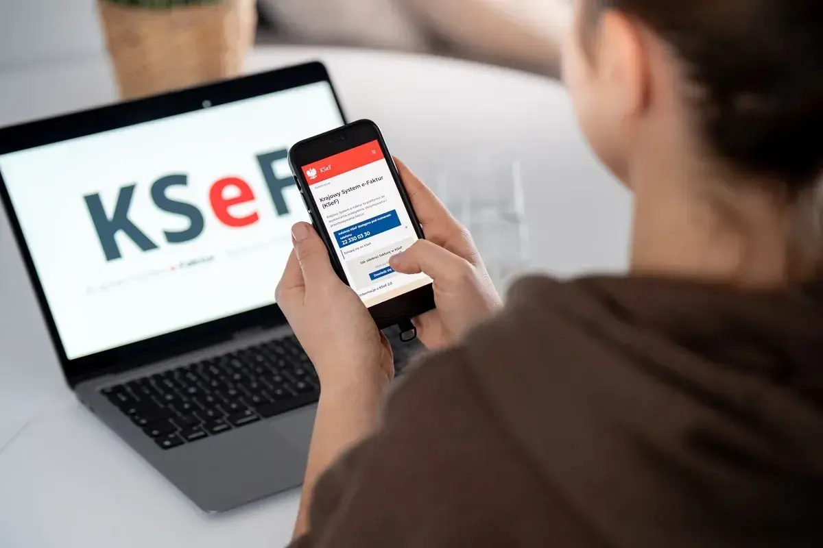 Osoba korzysta z telefonu z aplikacją KSeF, gdzie widnieje informacja o fakturze ustrukturyzowanej. Obok laptop z logo KSeF.