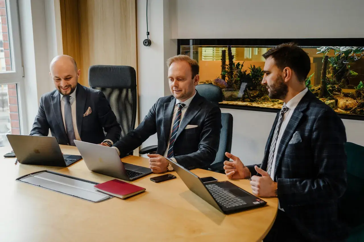 Trzech mężczyzn w garniturach przy drewnianym stole z laptopami i akwariem w tle.