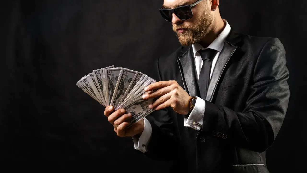 Elegancki mężczyzna w garniturze i okularach przeciwsłonecznych liczy banknoty. Wygląda jak windykator, który właśnie odzyskał dług.