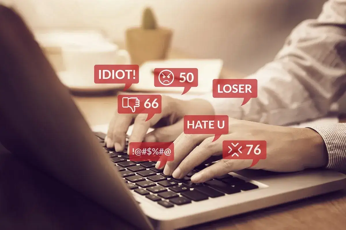 Dłonie piszą na klawiaturze, otoczone dymkami z obraźliwymi komentarzami, symbolizując zniesławienie w internecie.