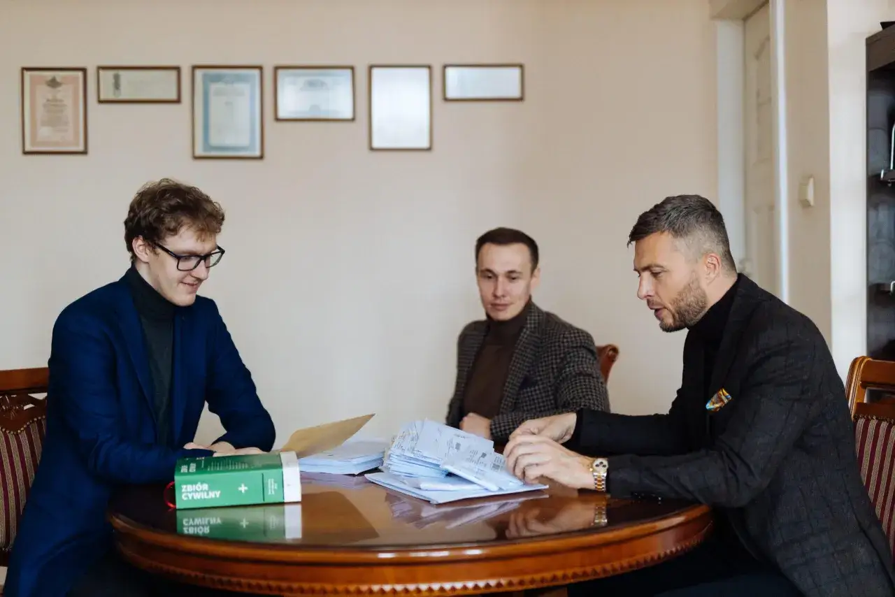 Trzech mężczyzn przy okrągłym stole omawia dokumenty. Jeden z nich, w garniturze, pokazuje stos papierów, zastanawiając się, ile bierze adwokat za reprezentowanie w sądzie.