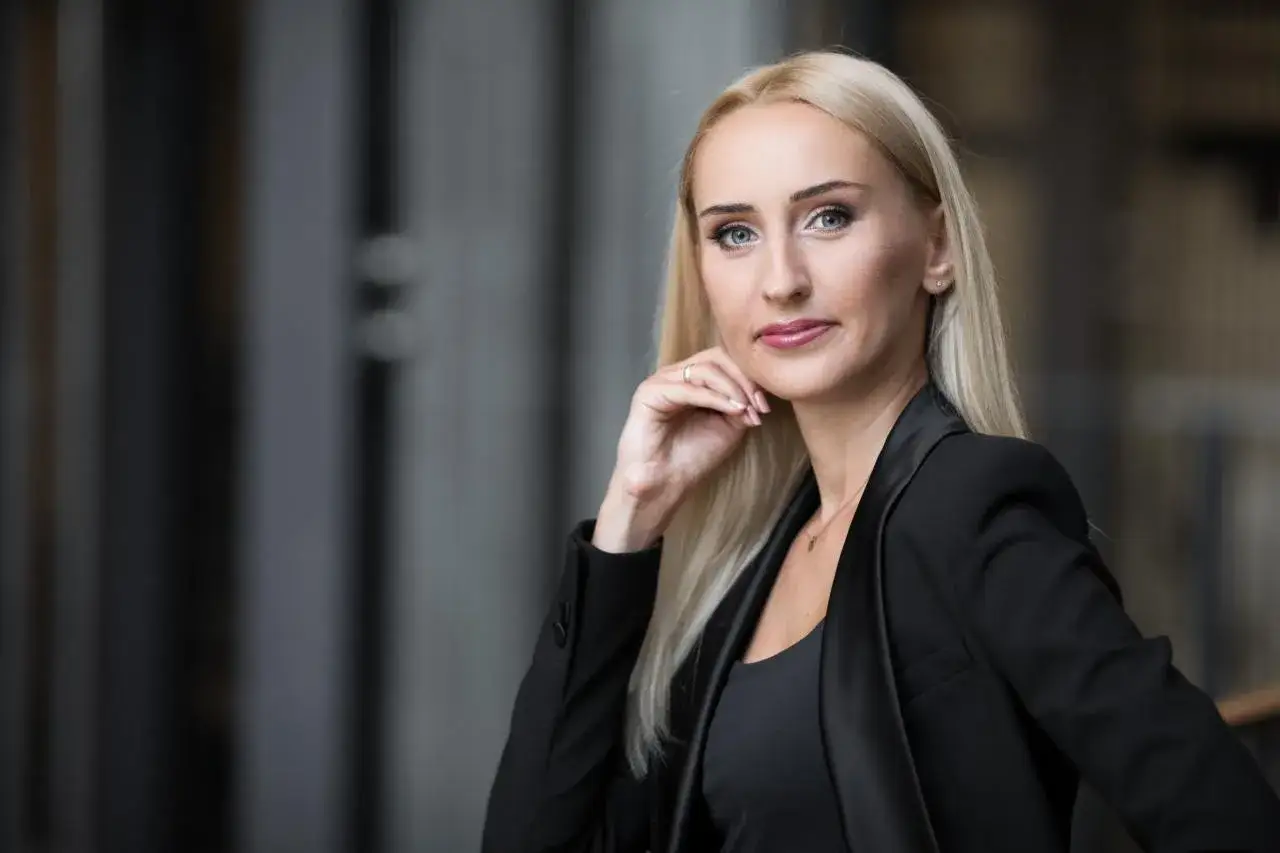 Portret kobiety z blond włosami w czarnej marynarce, opierającej podbródek na dłoni.
