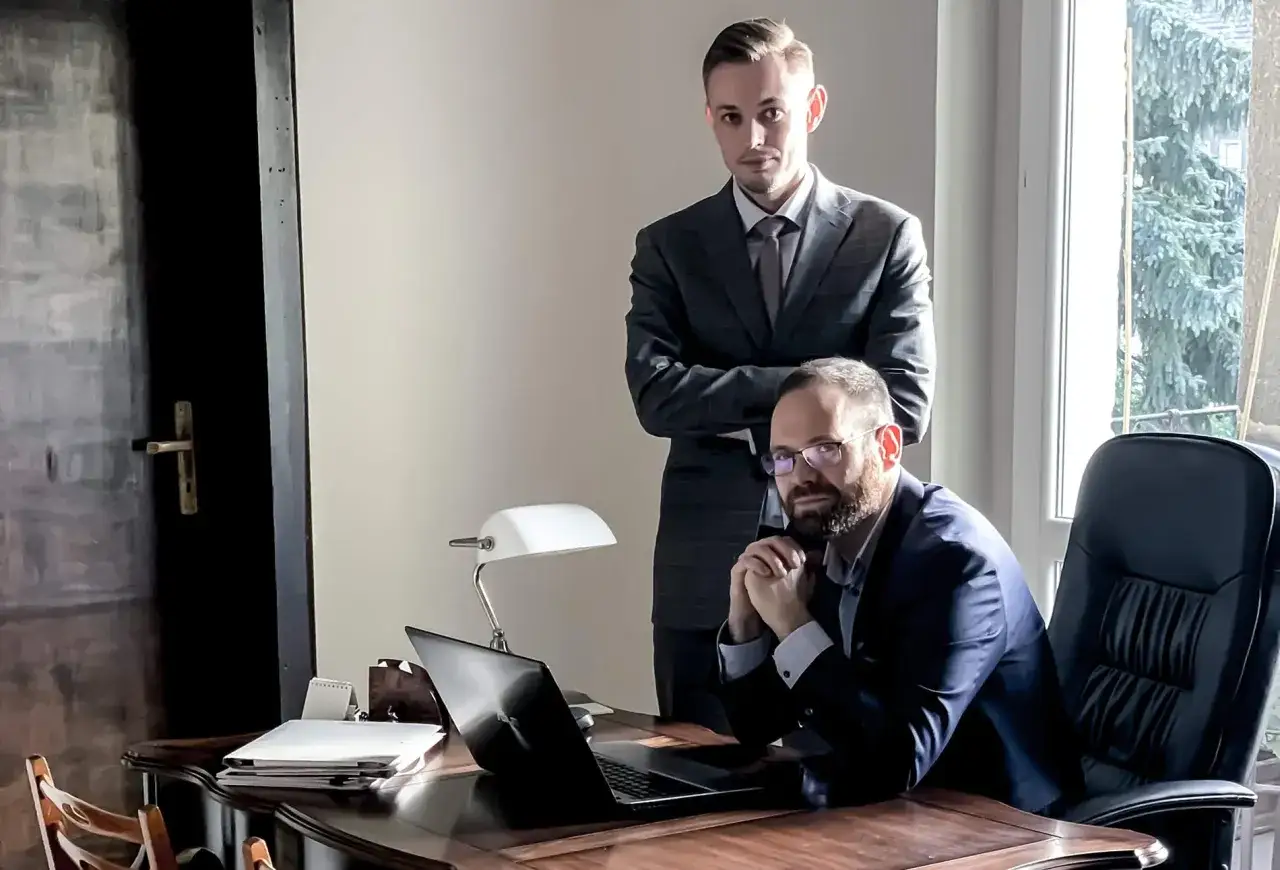 Dwóch mężczyzn w garniturach w biurze, jeden siedzi przy biurku z laptopem, drugi stoi za nim.