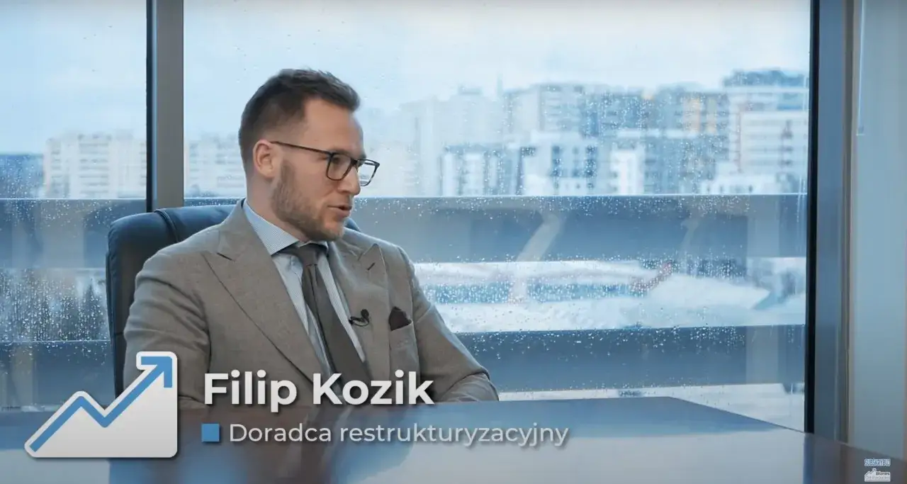 Mężczyzna w garniturze siedzi przy biurku, za nim okno z kroplami deszczu i widokiem na miasto.