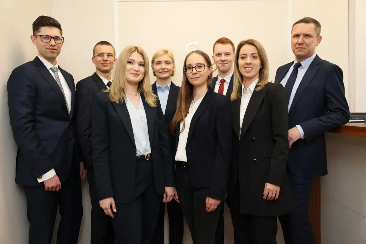 Grupa prawników w garniturach i kostiumach stoi przed jasnym tłem.