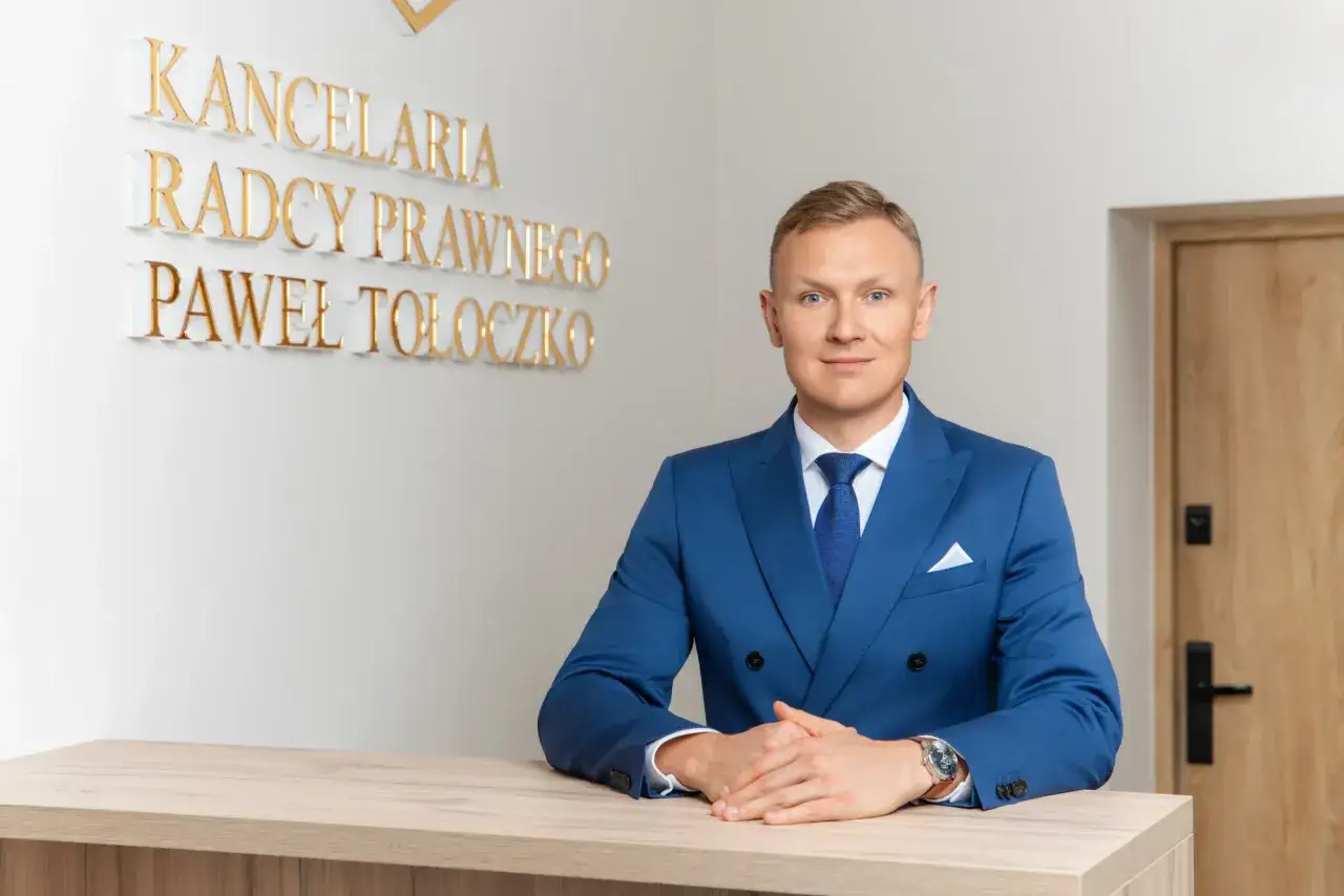 Mężczyzna w niebieskim garniturze przy biurku, za nim napis