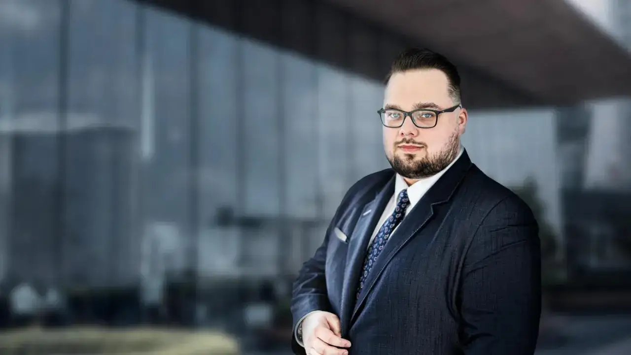 Mężczyzna w garniturze i okularach, na tle nowoczesnego budynku.