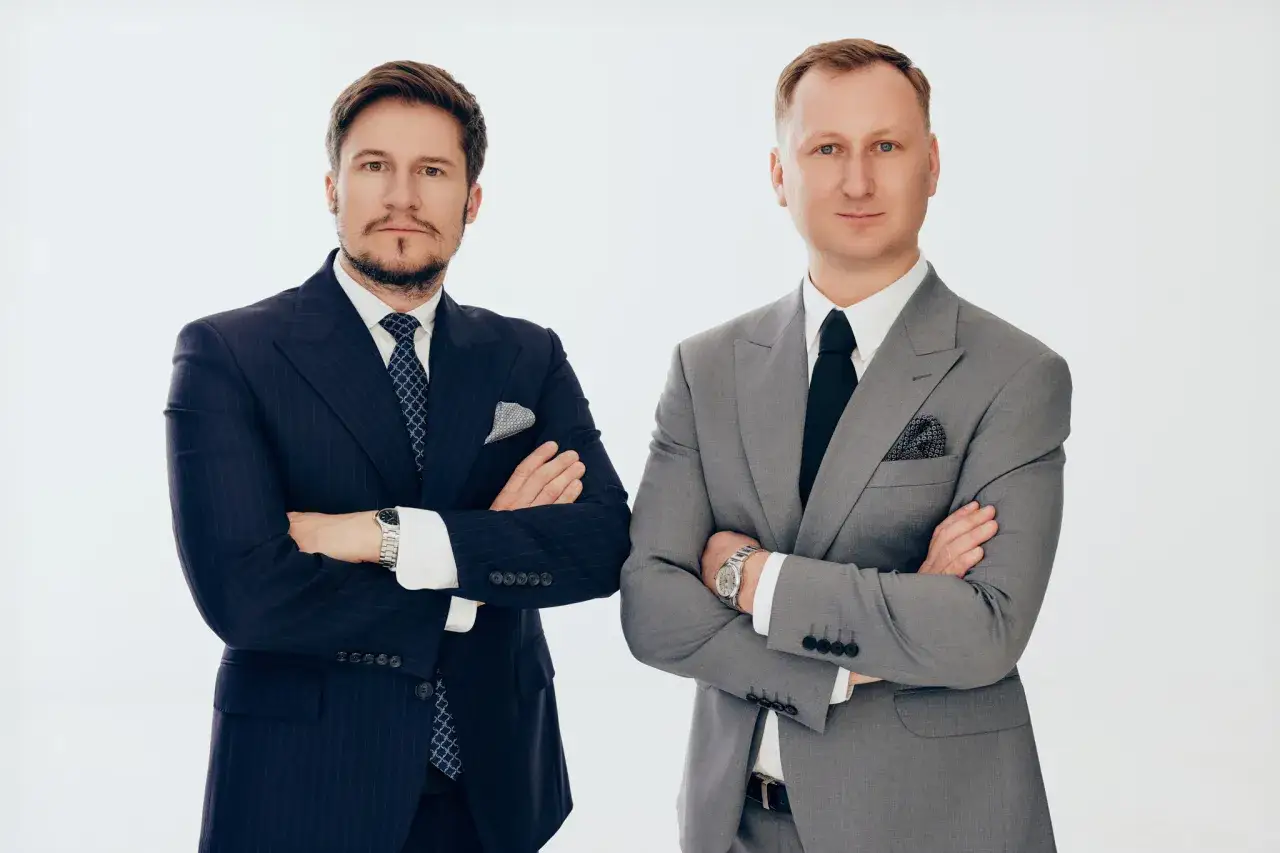 Dwóch mężczyzn w garniturach stoi ze skrzyżowanymi rękami na białym tle.
