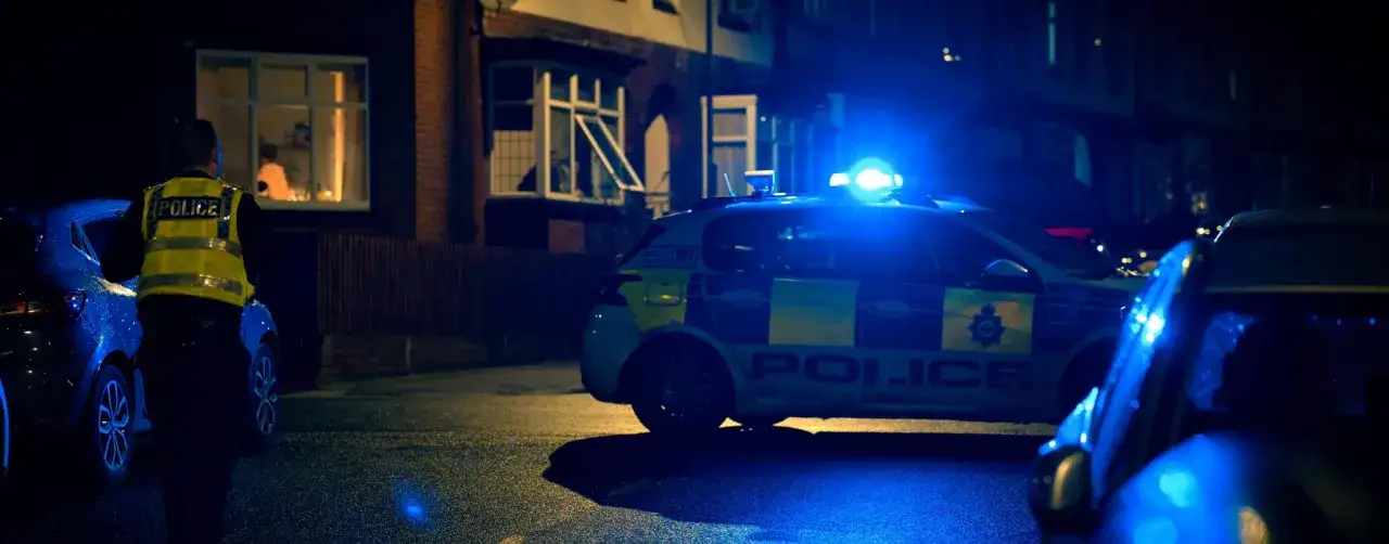 Nocna interwencja policji. Mundurowy w kamizelce odblaskowej stoi obok radiowozu z włączonymi sygnałami świetlnymi. Sytuacja może dotyczyć przemocy domowej.