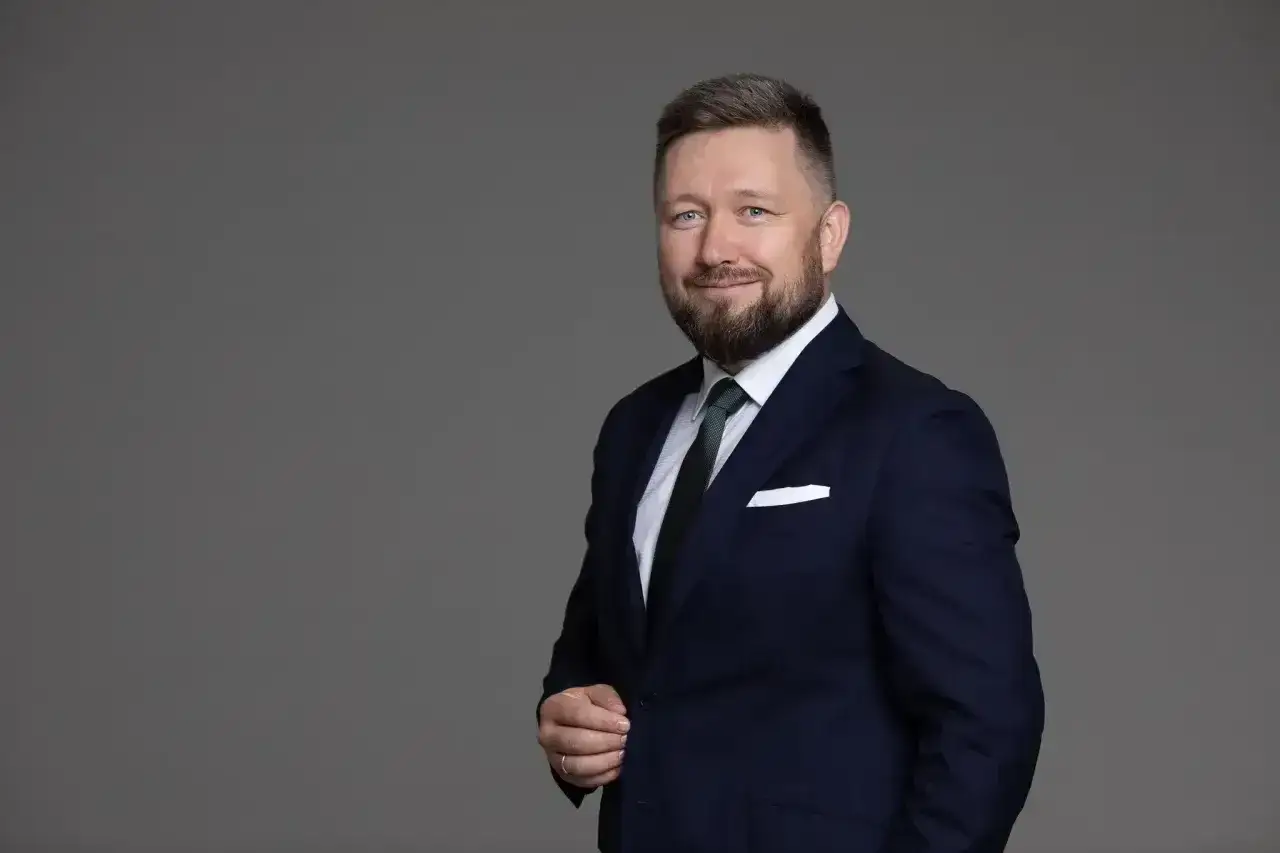 Mężczyzna w granatowym garniturze i krawacie, z brodą, uśmiecha się na szarym tle.