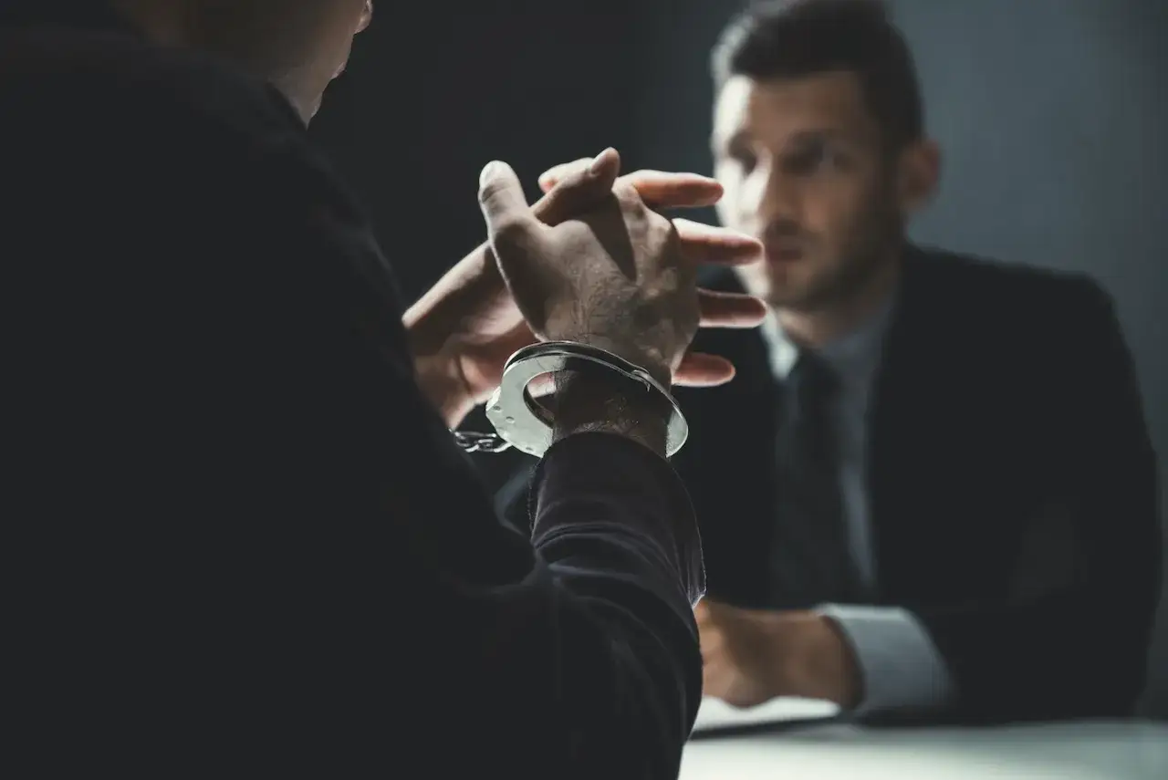 Dłoń w kajdankach na tle drugiego mężczyzny w garniturze.