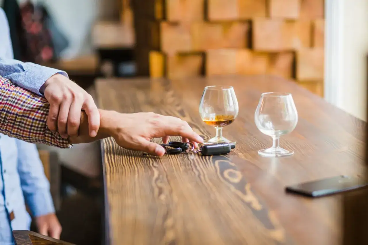 Ręka powstrzymuje drugą przed sięgnięciem po kluczyki do samochodu. Obok kieliszek z alkoholem. Zakaz prowadzenia pojazdów po alkoholu.