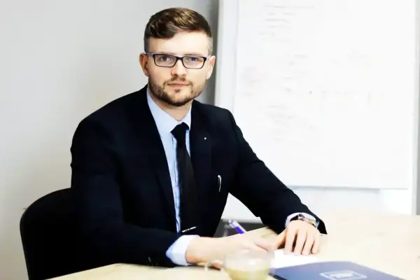 Mężczyzna w garniturze i okularach siedzi przy biurku, pisząc coś na kartce. Obok stoi filiżanka kawy.
