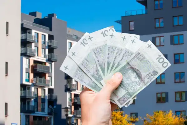 Dłoń trzyma banknoty 100 zł na tle nowoczesnych bloków mieszkalnych. To symbol ulgi mieszkaniowej i możliwości zakupu własnego M.