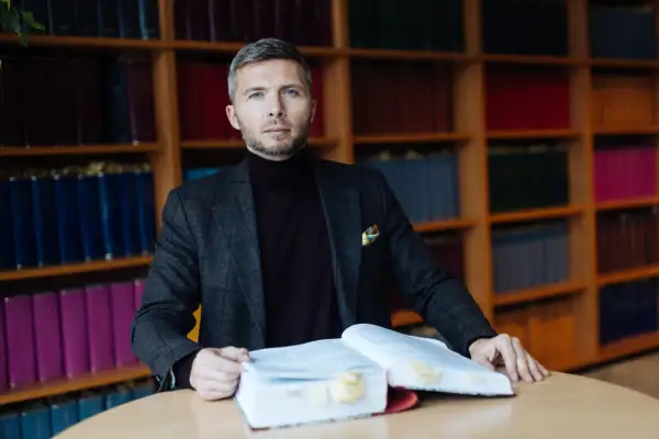 Mężczyzna w garniturze siedzi przy stole z otwartą księgą, otoczony regałami z książkami. Zastanawia się, ile bierze adwokat za reprezentowanie w sądzie.