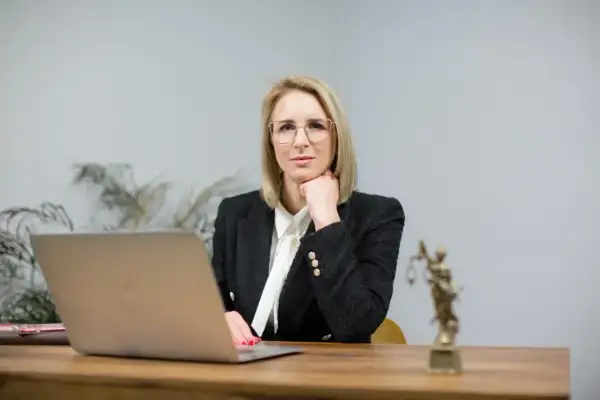 Kobieta w okularach i żakiecie siedzi przy biurku z laptopem i Statuą Temidy.