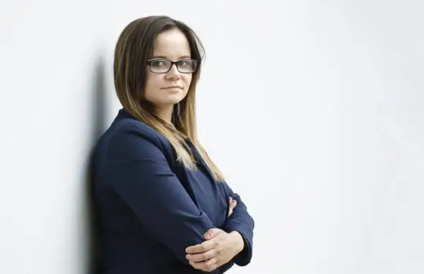 Kobieta w okularach i granatowej marynarce, oparta o białą ścianę.