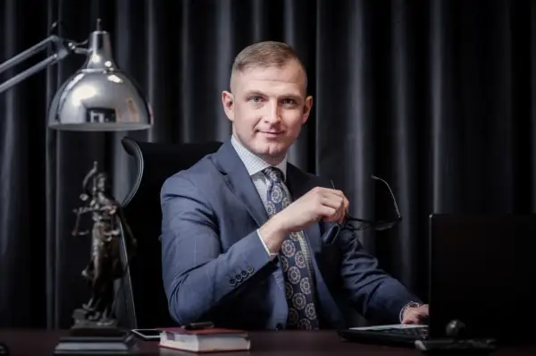 Mężczyzna w garniturze siedzi przy biurku, trzymając okulary. Obok statua Temidy.